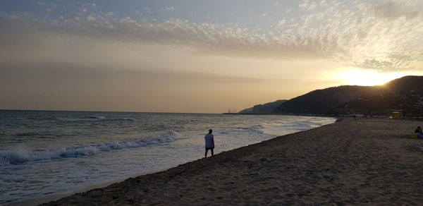 Rear view of man walking at beach against sky during sunset