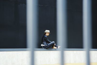 Side view of woman sitting on retaining wall seen through fence