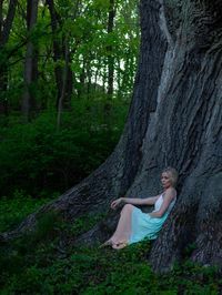 Full length of woman sitting by tree trunk in forest