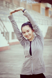 Portrait of smiling young woman stretching while standing against built structure