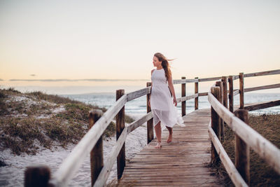 Young woman walking on pier at beach against clear sky during sunset