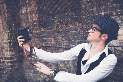 Mature man photographing while standing against brick wall during autumn