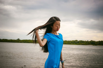 Young woman standing at riverbank against cloudy sky