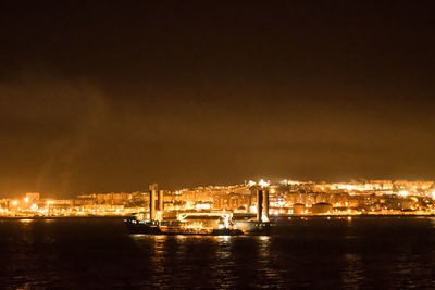 Illuminated buildings by sea against sky at night