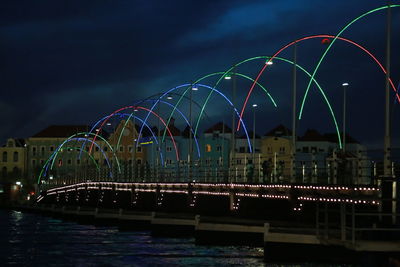 Illuminated colorful bridge over river against cloudy sky at night