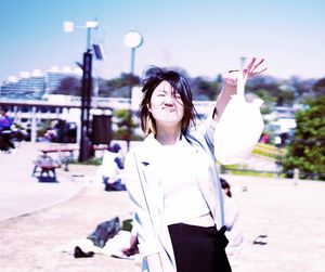 Portrait of smiling young woman holding plastic bag while making a face against sky