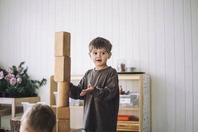 Boy looking away while standing near tower of toy blocks in classroom at kindergarten