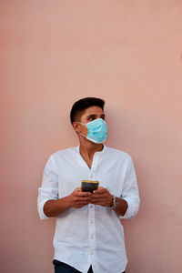 Young latin man with mask is looking his phone on pink background