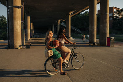 Side view of teenage girl sitting with friend riding bicycle on road during sunny day