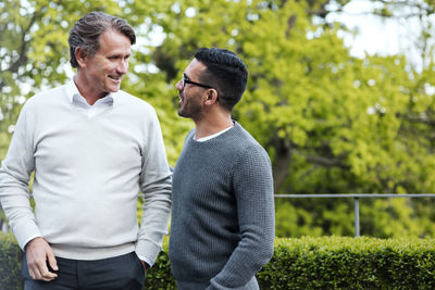 Smiling multi-ethnic friends talking while standing against hedge in yard