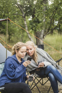 Mother and daughter looking at smart phone while having coffee in campsite