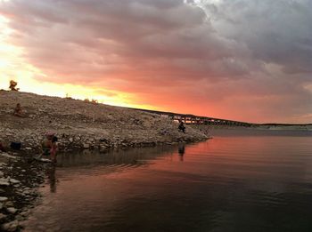 People by lake against cloudy sky during sunset