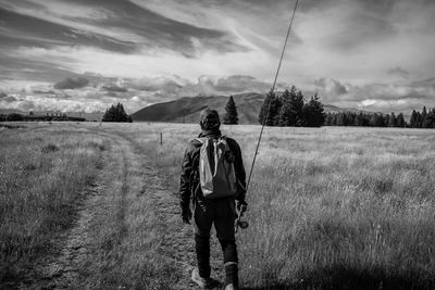 Rear view of man with fishing rod walking on field against sky