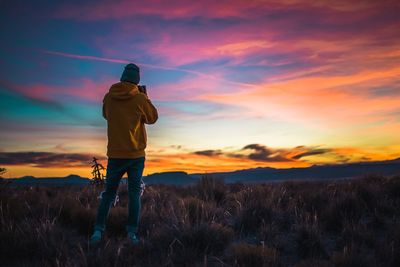 Rear view of man photographing while standing on field against sky during sunset