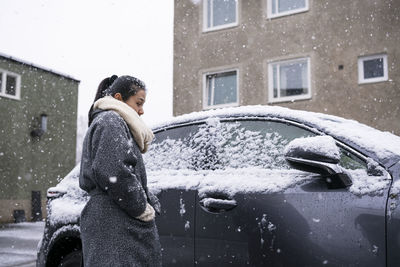 Woman standing on snow covered car against building during winter