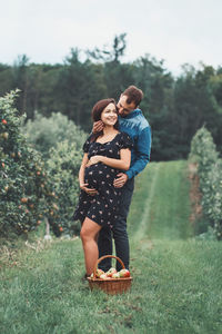 Full length of pregnant couple embracing by apples in basket on grass