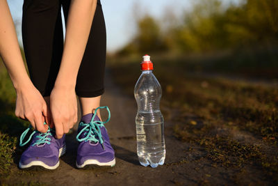 Cropped image of woman tying shoelace with water bottle on field