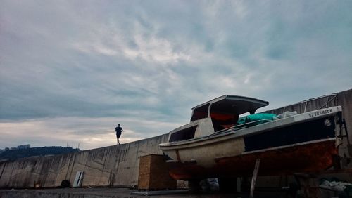 Low angle view of man running on retaining wall by boat moored on pier against cloudy sky
