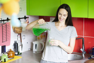 Beautiful young woman pouring green juice drink in the glass at home kitchen