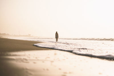 Rear view of woman standing on shore at beach against clear sky during sunset