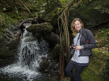 Portrait of confident woman holding camera while standing by waterfall in forest