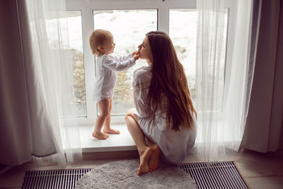 Mother in a white robe sits with a child a blonde daughter at a large window of the house person