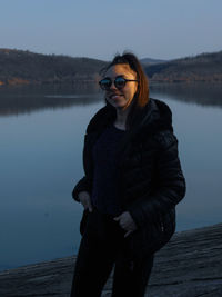 Young woman wearing sunglasses standing by lake against sky