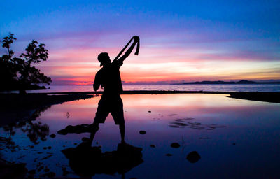 Silhouette man holding belt while standing at beach against sky at dusk