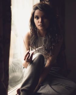 Portrait of young woman holding eyeglasses while sitting on window sill