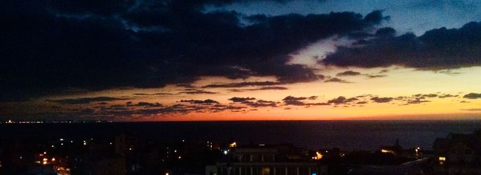 Silhouette cityscape by sea against dramatic sky