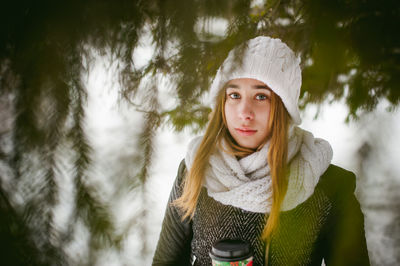 Young woman wearing warm clothing while holding disposable cup amidst trees during winter