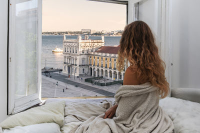 Rear view of young woman looking at view through window while relaxing on bed in hotel