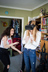 Cheerful teenage girl looking at friend wearing virtual reality headset at home