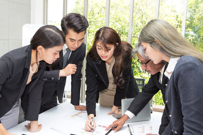 Colleagues discussing data on desk while standing against window in office