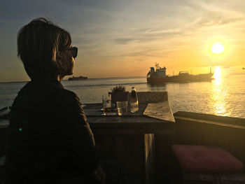 Rear view of woman sitting by sea against sky during sunset