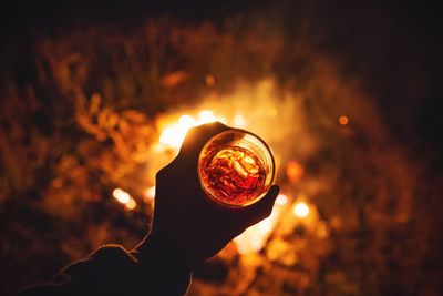 Cropped hand of person holding drink in glass over bonfire