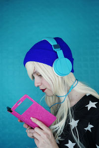 Close-up of woman with blond hair listening music while using mobile phone against blue wall