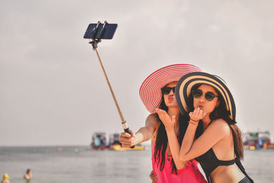 Female friends taking selfie with monopod at beach against sky