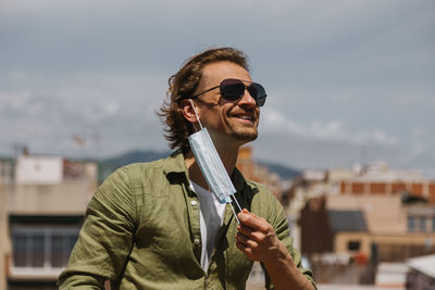 Handsome man in sunglasses joyfully removes the medical mask from his face on sunny day, outdoor. 