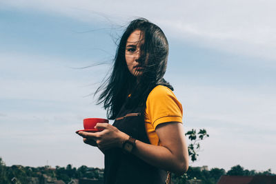 Young woman drinking drink standing against sky