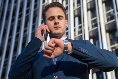 Low angle view of businessman talking on phone while checking time in city