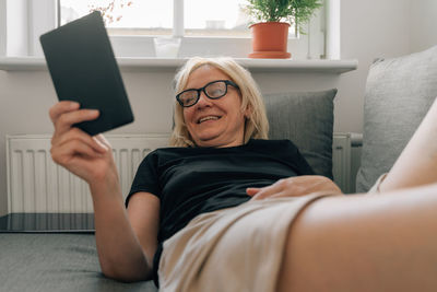 Smiling woman reading e-book by lying on sofa at home