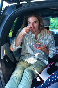 Young man sitting in car trunk and eating cake