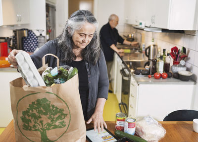 Senior woman removing grocery from shopping bag while using digital tablet in kitchen