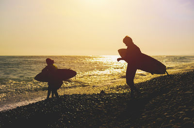 Silhouette father and son with surfboard at beach against sky during sunset. 