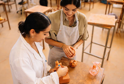 From above merry hispanic girlfriends smiling and creating geometric shape from clay on table in spacious workshop