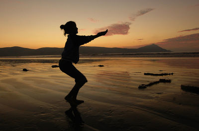 Silhouette young woman dancing at beach against sky during sunset