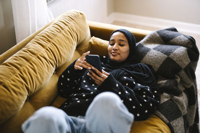 Smiling young woman using smart phone lying on sofa at home