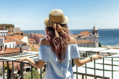 Rear view of young woman looking at buildings and sea while standing by railing in balcony against clear sky