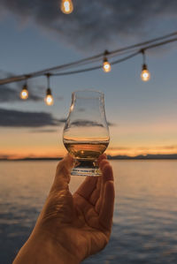 Close-up of hand holding wineglass against sea during sunset